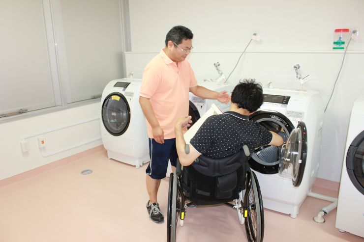機能訓練棟　洗濯室で車いす利用者が介護員に見守られてドラム式洗濯乾燥機で選択している写真