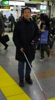 写真。男性が駅のコンコースを単独で移動している様子。比較的混雑している駅の中を、視覚障害者用誘導ブロックや白杖を活用。