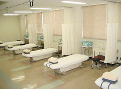 写真。臨床実習室の様子。施術用昇降ベッドが窓沿いに数台配置され、各ベットの間は、カーテンで仕切ることができる。ベッドサイドワゴン、キャスター付き丸椅子もベッドごとに配置されている。