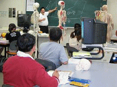 写真。授業風景。解剖学教室での授業の様子。利用者4人程で一つの大机を共有する。大机は4台あり、授業がわかり易いよう配慮して、各机に同じ解剖模型を配っている。この写真では、各机の上に頭蓋骨の模型がある。また、授業中に拡大読書器を使用する利用者の姿もある。