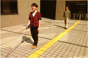 写真。歩行訓練の様子。夏、晴天。2人の人物が訓練中。利用者が、路面に設置された視覚障害者用誘導ブロックに沿って、白杖を用いて歩いている。その後ろから、指導員が、利用者の様子を確認しつつ歩いている。