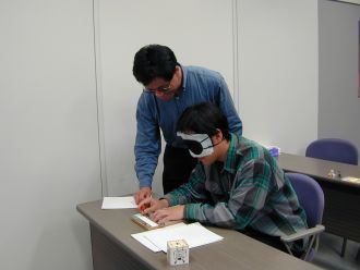 画像です。アイマスクをした男性が点字を打つのを男性の指導員が指導しています