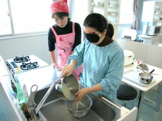 画像です。アイマスクをした女性がお米を研ぐ様子を女性の指導員がかたわらで見守っています