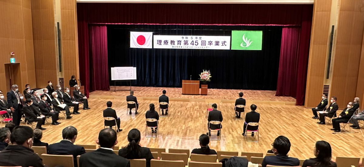 令和5年度理療教育第45回卒業式 卒業式の写真、本館1階講堂にて。8名の卒業生がフロア側中央付近に設置した椅子に着席、右手にご来賓、左手に関係者が臨席。