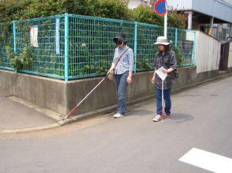画像です。アイマスクをして白杖を持った女性が住宅街の角を曲がろうとする様子を女性の指導員がかたわらで見守っています