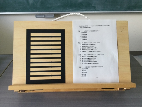 視覚支援用具 （左側）罫プレート。手紙や文字を書くときに使います。 （右側）試験問題 （後方）木製の書見台。角度を調整することで、体へ負担が少ない自然な姿勢で文字を見ることができます。