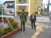 写真。歩行訓練の様子。市街地の交差点で、男性の利用者と指導員が、訓練に取り組んでいる様子。