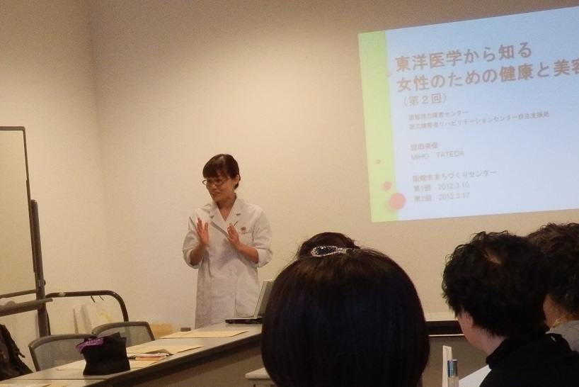 写真。講座の様子。白衣を着た女性職員が、講義をしているところ。正面のスクリーンにスライドが映し出されている。演題「東洋医学から知る、女性のための健康と美容」