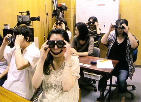 写真。弱視体験用のレンズをかけている受講者の皆さん。座席に座った５人の女性が、それぞれ、体験レンズを使って弱視者の見え方を確認している。その中央に、女性の支援員がおり、説明や助言をしている。