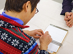 写真。点字訓練の様子。利用者が標準点字盤を使い、点字を書いているところ。