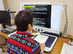 写真。ロービジョン訓練。利用者が拡大読書器を使い、ディスプレイを白黒反転画面にし、拡大文字を見ている。